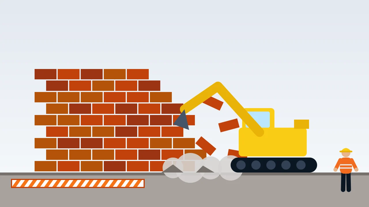 An excavator knocking down a brick wall behind a safety barrier